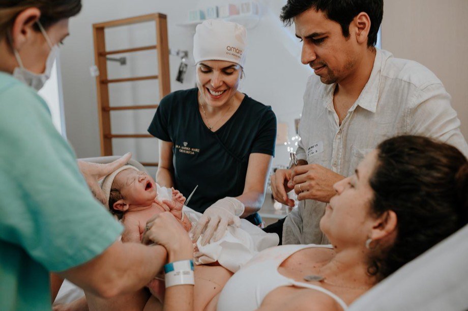 Médica obstetra segura o recém-nascido logo após o parto enquanto o pai corta o cordão umbilical e a mãe acompanha da cama.