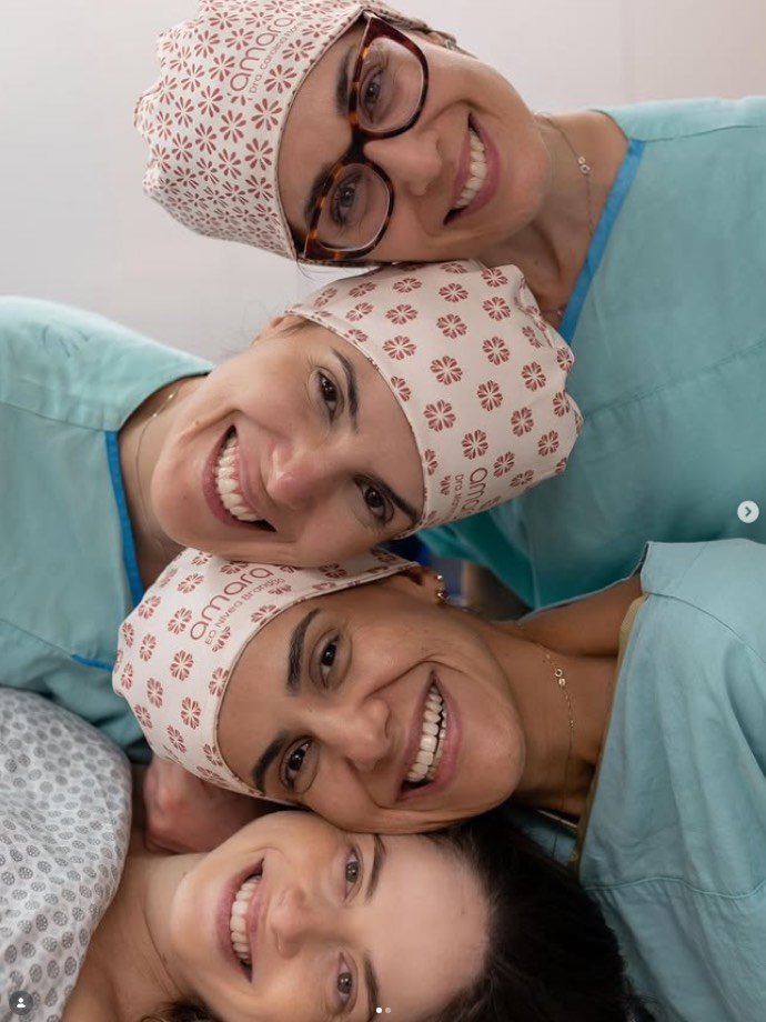 Quatro profissionais de saúde com toucas cirúrgicas iguais e pijamas verdes sorriem juntas, com os rostos encostados.