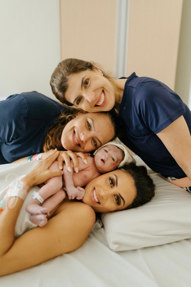 Mãe, recém-nascido e duas profissionais de saúde deitados juntos na cama do quarto, com os rostos encostados e sorrindo.